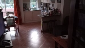 Une cuisine avec des meubles blancs, une table à manger avec des chaises, un téléviseur posé sur un meuble et une fenêtre avec des rideaux.