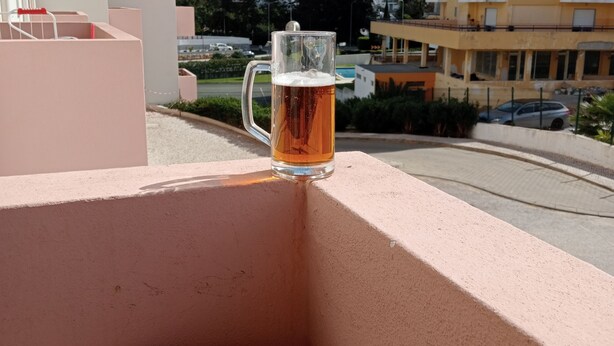 Un verre de bière sur un balcon, avec vue sur un immeuble de grande hauteur et un bâtiment jaune en arrière-plan.