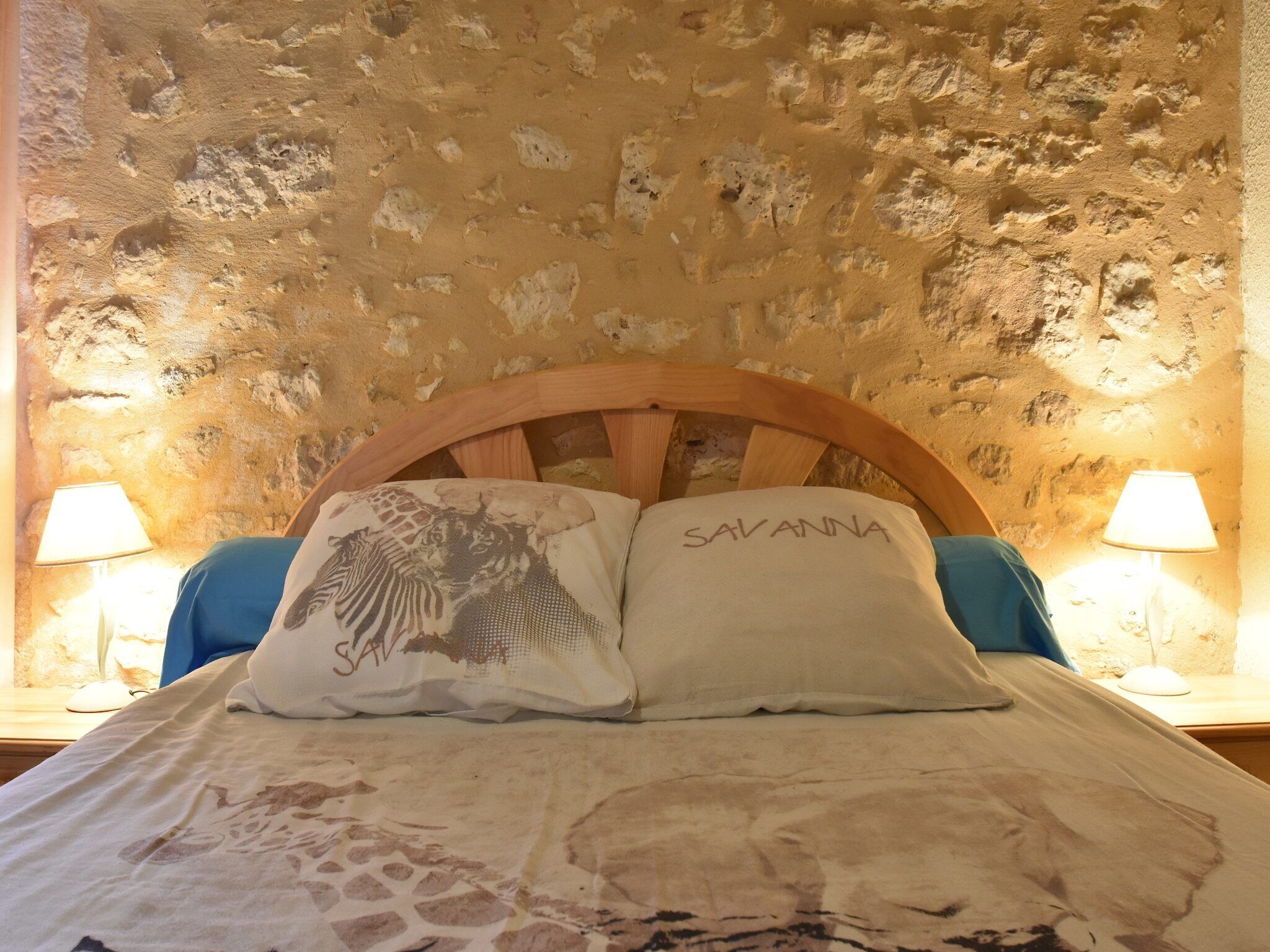 Un lit avec deux oreillers, l’un orné d’un motif girafe et l’autre portant le nom « SAVANNA ». La chambre est dotée d’un mur en pierre et de deux lampes de chevet.