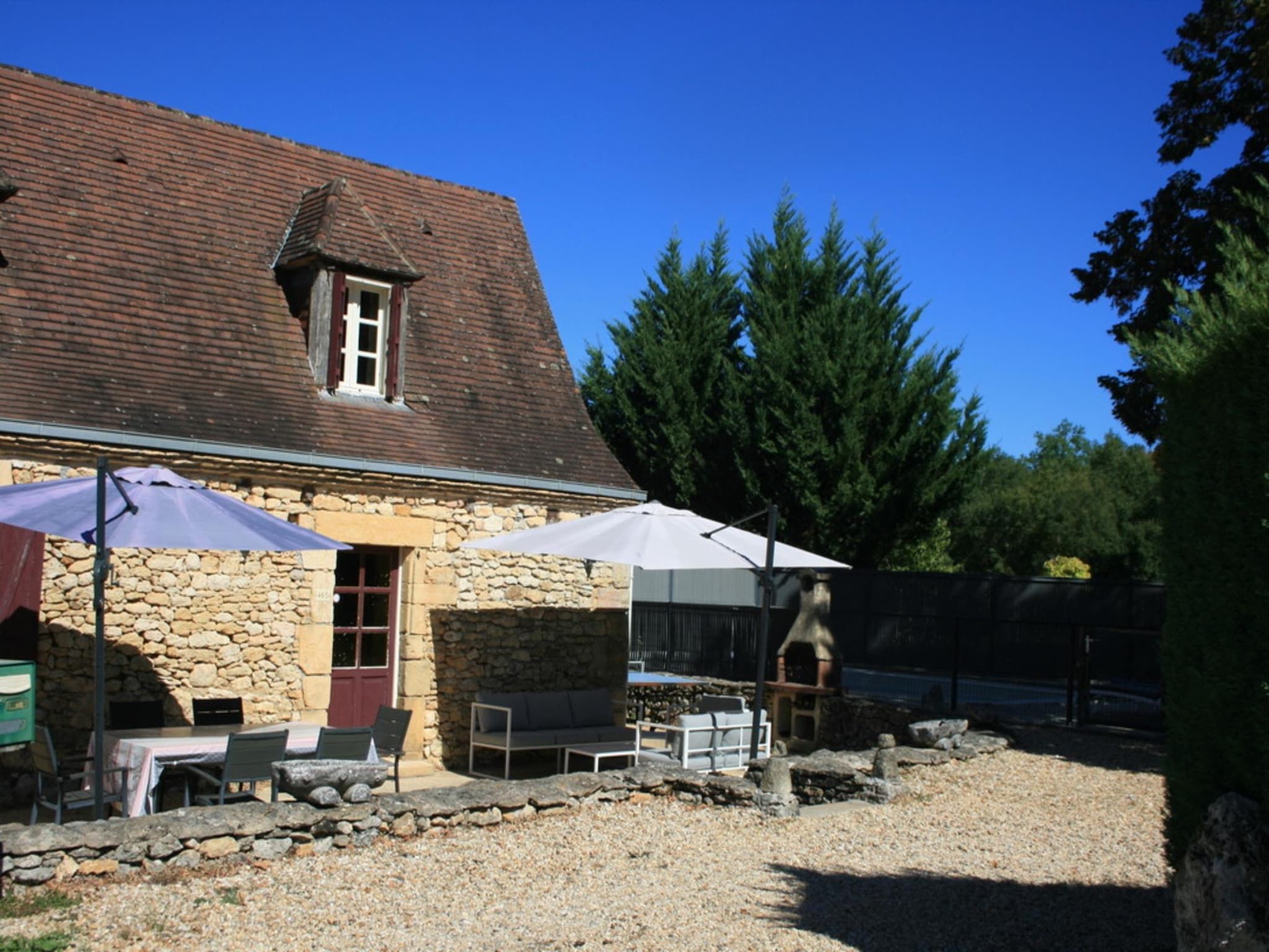 A stone building with a tiled roof, outdoor seating with umbrellas, and a large tree in the background.