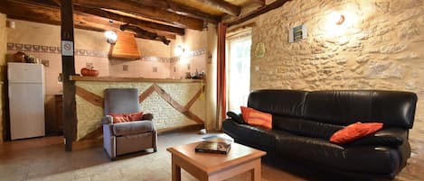 A cozy living room with a stone wall, wooden ceiling beams, a black leather sofa, a wooden coffee table, and a patterned armchair.