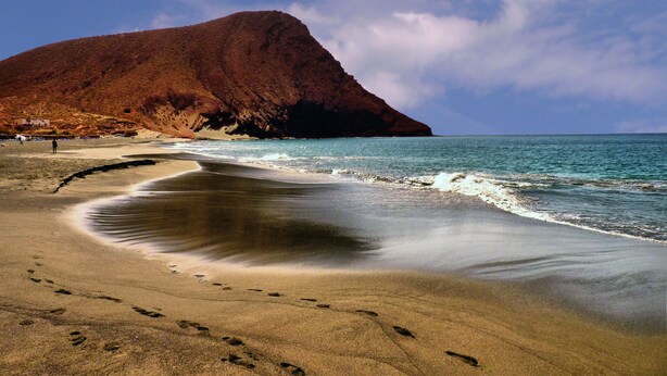 Ein Strand mit Fußspuren, die zum Wasser führen, ein großer Berg im Hintergrund und ein klarer Himmel.
