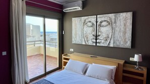 Ein Hotelzimmer mit einem Bett, einem Nachttisch, einer Lampe, einem großen Fenster mit Vorhängen und einer Wandklimaanlage.
