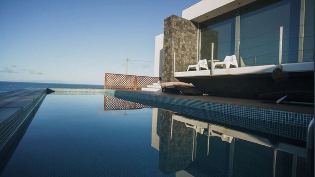 Ein moderner Infinity-Pool mit freiem Blick auf den Ozean, einer Steinsäule und einer Lounge-Bereich mit weißen Stühlen.