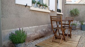 A wooden deck with two chairs and a table, potted plants, and a window with shutters.