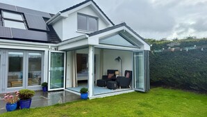 A modern house with a glass-enclosed patio area, a visible bedroom with a bed, and a well-maintained lawn. - Luxury Riverside Holiday Home in stunning Ballina / Killaloe - The Derg House (Killaloe)