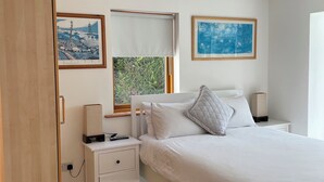 A bedroom with a bed, bedside tables, a nightstand, a framed picture, and a window with a view of greenery. - Luxury Riverside Holiday Home in stunning Ballina / Killaloe - The Derg House (Killaloe)