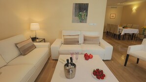A modern living room with a sofa, a coffee table with bottles and strawberries, and a dining area in the background.