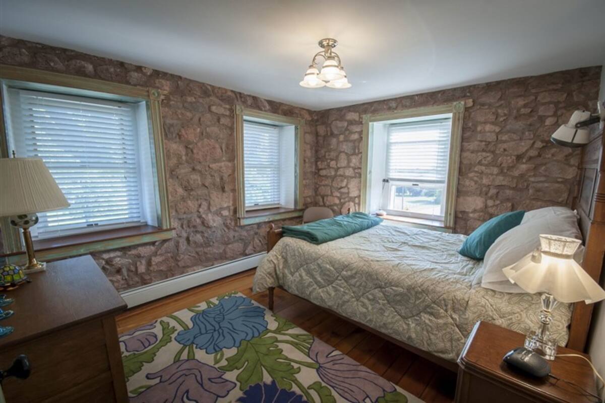 A bedroom with a stone wall, a bed with a quilt, a nightstand, a lamp, and a window with blinds.