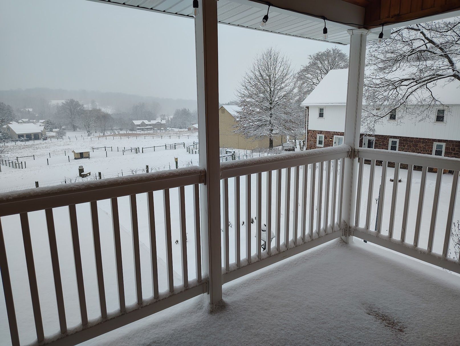 A snow-covered balcony with a view of a rural landscape and a building with a white roof.