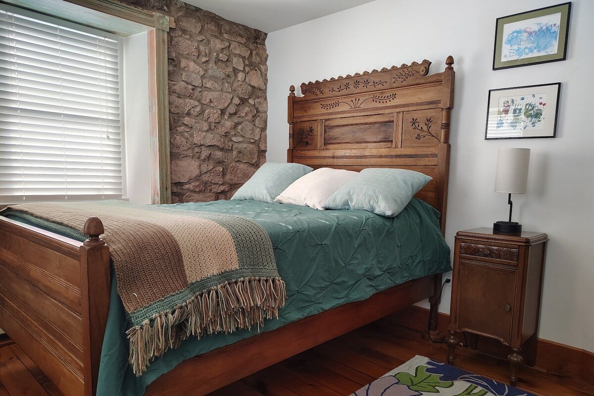 Quarto com cama de madeira, parede de pedra e mesa de cabeceira com abajur.