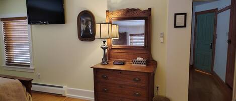 A bedroom with a wooden dresser, a television mounted on the wall, a bedside table with a lamp, and a window with blinds.