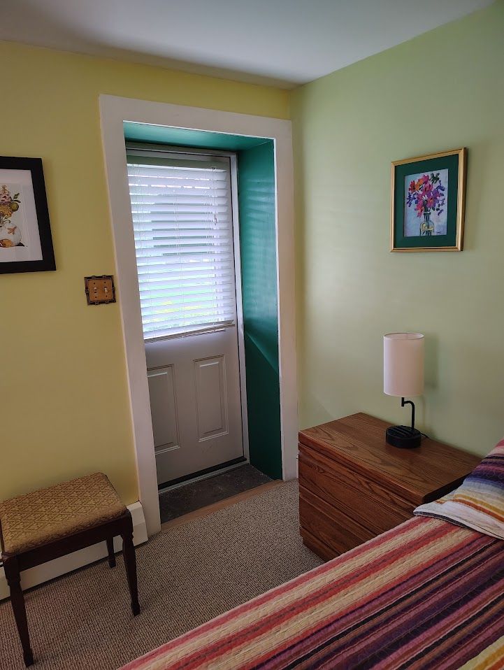 A bedroom with a bed, a bedside table with a lamp, a chair, a door with a window, and a framed picture on the wall.