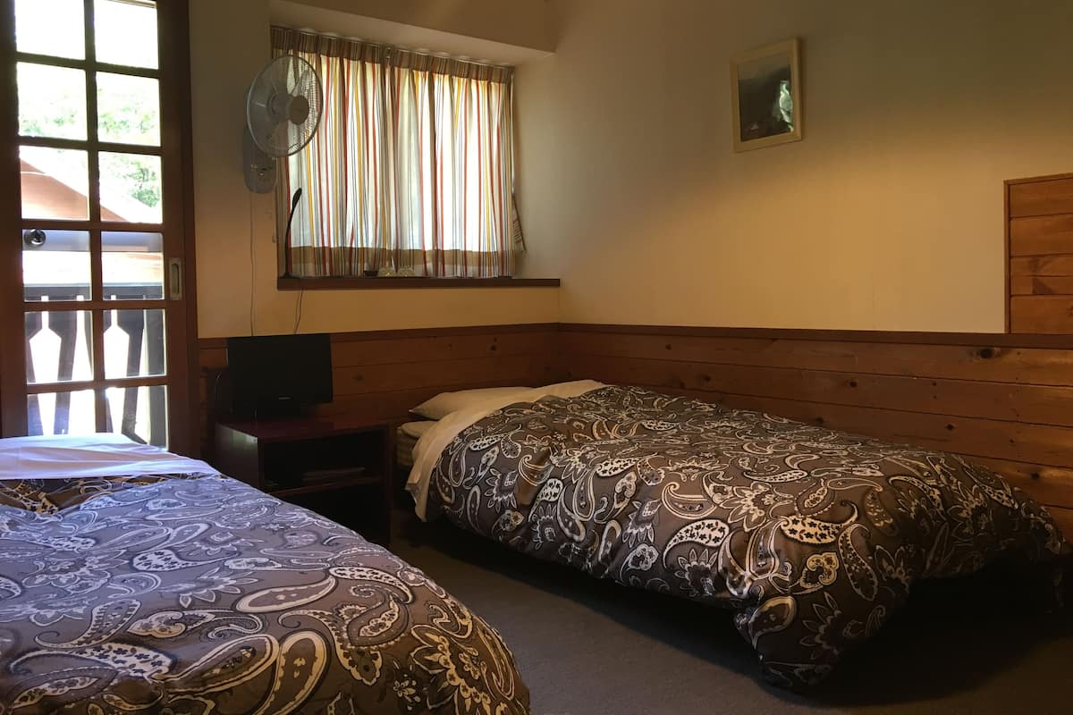 A room with two beds, a window with curtains, a fan, a painting, and a wooden headboard.