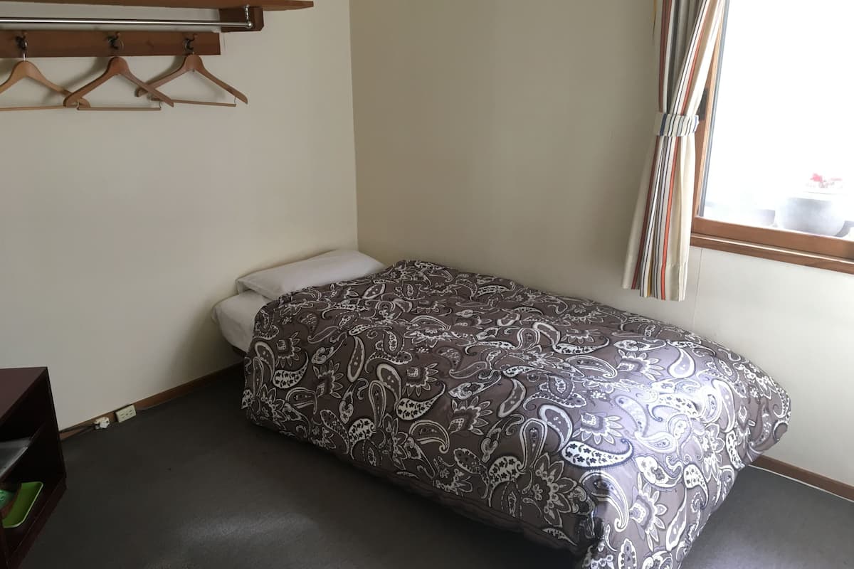 A small, single-bed room with a patterned bedspread, a wooden coat rack, and a window with curtains.