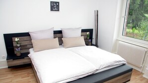 Ein modernes Schlafzimmer mit einem Holz-Kopfteil, einem großen Bett mit weißer Bettwäsche, einem kleinen Nachttisch mit Pflanze und Blick auf Grün durch ein Fenster.