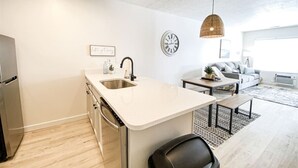 Une cuisine moderne dotée d’un îlot central, un coin repas avec un canapé et une table, et un salon avec un divan et une grande fenêtre.