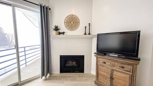 Un salon avec une cheminée, un téléviseur posé sur un meuble en bois et une grande fenêtre avec des rideaux.