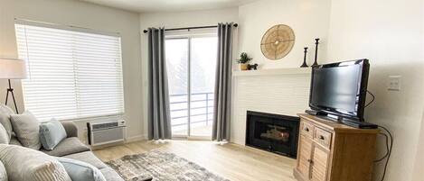 A living room with a sofa, a television on a wooden cabinet, a fireplace, and a large window with curtains.