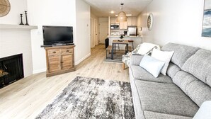 Un salon avec un canapé gris, un téléviseur posé sur un meuble en bois, une cheminée et, au fond, une cuisine.
