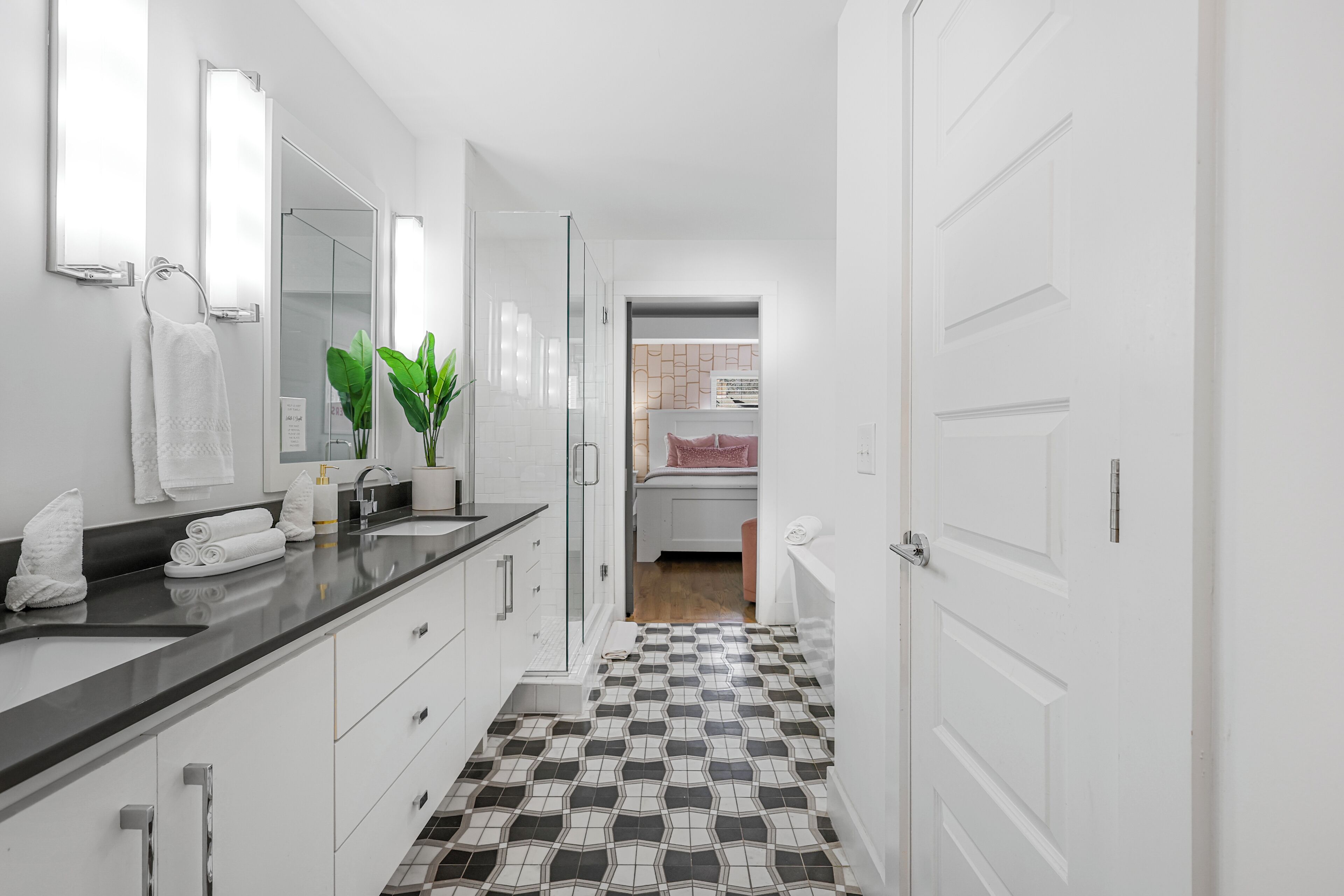 Een moderne badkamer met een zwart aanrechtblad, witte kasten, een glazen douchecabine en een vloer met een patroon.