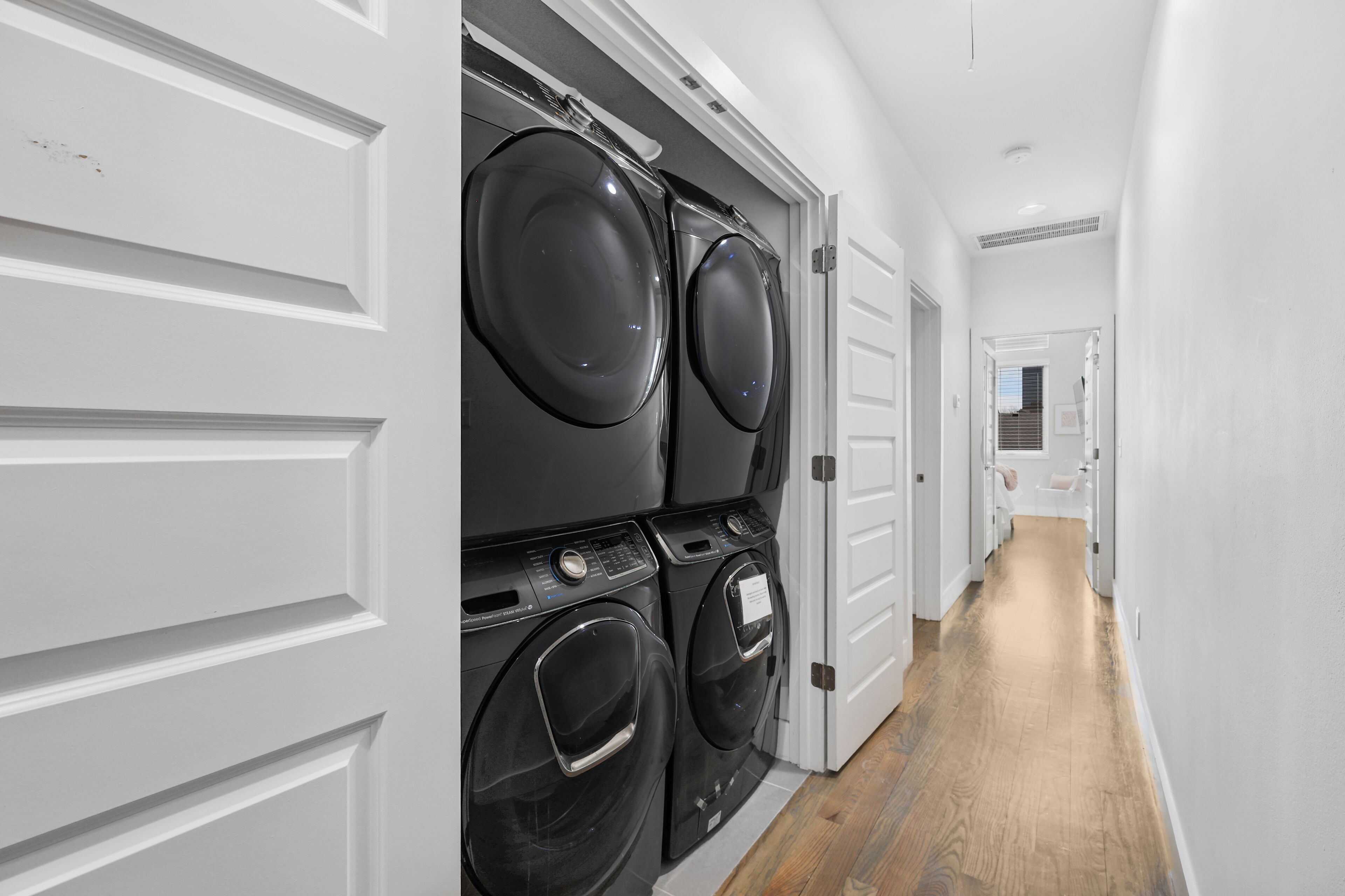 A laundry room with two stacked front-loading washing machines, a white door, and a wooden floor.