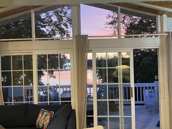 Une terrasse couverte offrant une vue sur un lac au coucher du soleil, un canapé noir orné d’un coussin à motifs de poissons et une rambarde en bois.