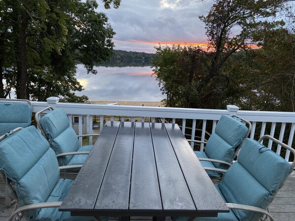 Une terrasse en bois avec une table et des chaises, offrant une vue sur un lac paisible et une rive lointaine.