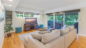 Een moderne woonkamer met een grote flatscreen televisie, een beige bank en een houten salontafel. In de hoek staat een potplant en een boekenkast.
