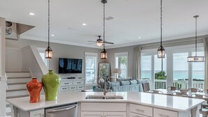 A modern kitchen with white cabinetry, a large island, and pendant lighting. There is a dining area with a view of the ocean.