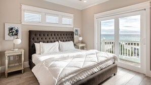 A bedroom with a large bed, a headboard, a nightstand, a lamp, and a view of the ocean through a sliding door.