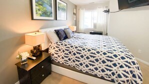 Een slaapkamer met een bed, nachtkastje, lamp en een aan de muur bevestigde televisie.