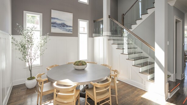 A modern interior with a round dining table, wooden chairs, a potted plant, and a staircase with glass railings.