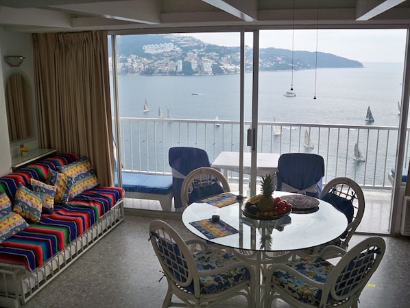 A balcony with a sofa and chairs, a table with fruit, and a view of the sea and buildings.