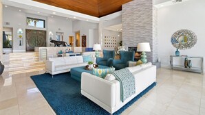 A spacious living area with a white sofa, teal armchairs, and a blue rug. A large window provides natural light, and the room features a wooden ceiling and a textured wall. - Newly Remodeled 4,400 Square Foot Modern design with Pool and Spa. #065893 (La Quinta)