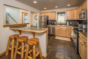 A kitchen with a central island, wooden stools, granite countertops, stainless steel appliances, and wooden cabinets.