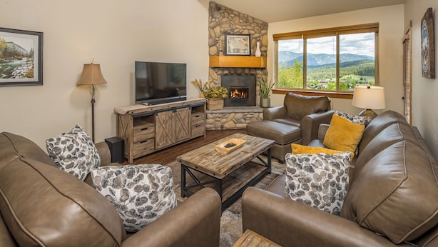 Ein Wohnzimmer mit Ledersofas, einem Kamin aus Stein, einem Fernseher und einem großen Fenster mit Blick auf die Berge.