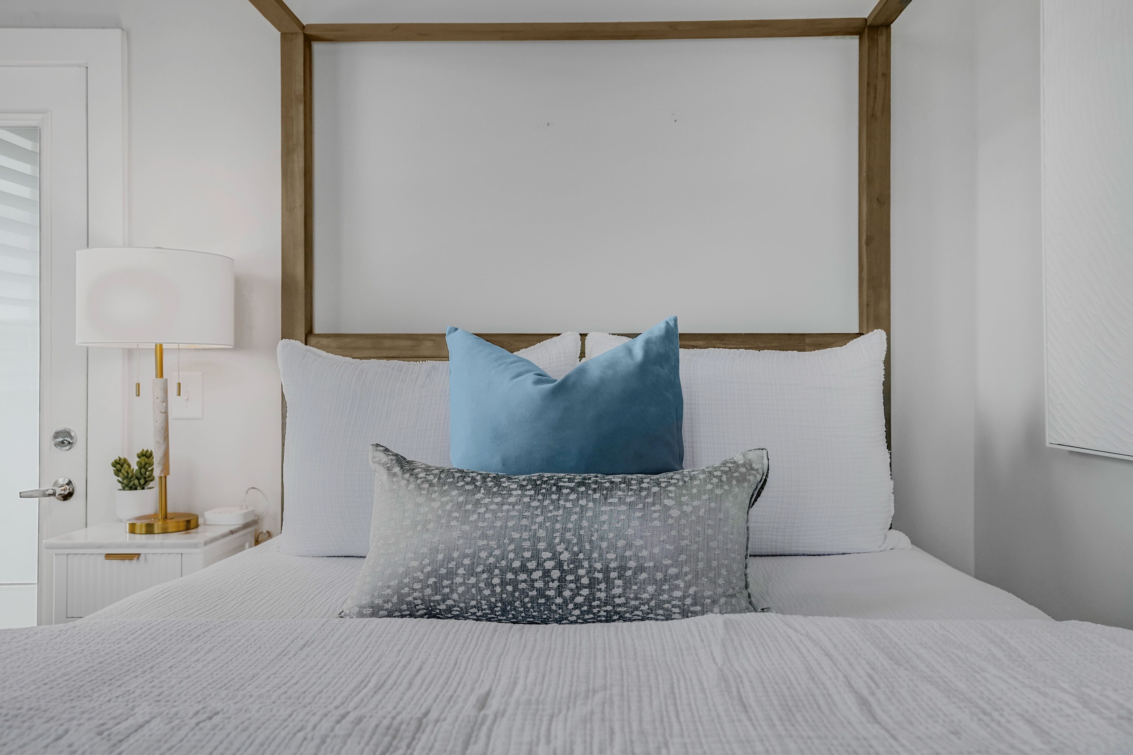 A neatly made bed with white bedding and two pillows, a bedside table with a lamp, and a small plant.