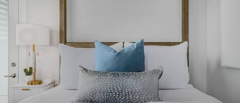 A neatly made bed with white bedding and two pillows, a bedside table with a lamp, and a small plant.