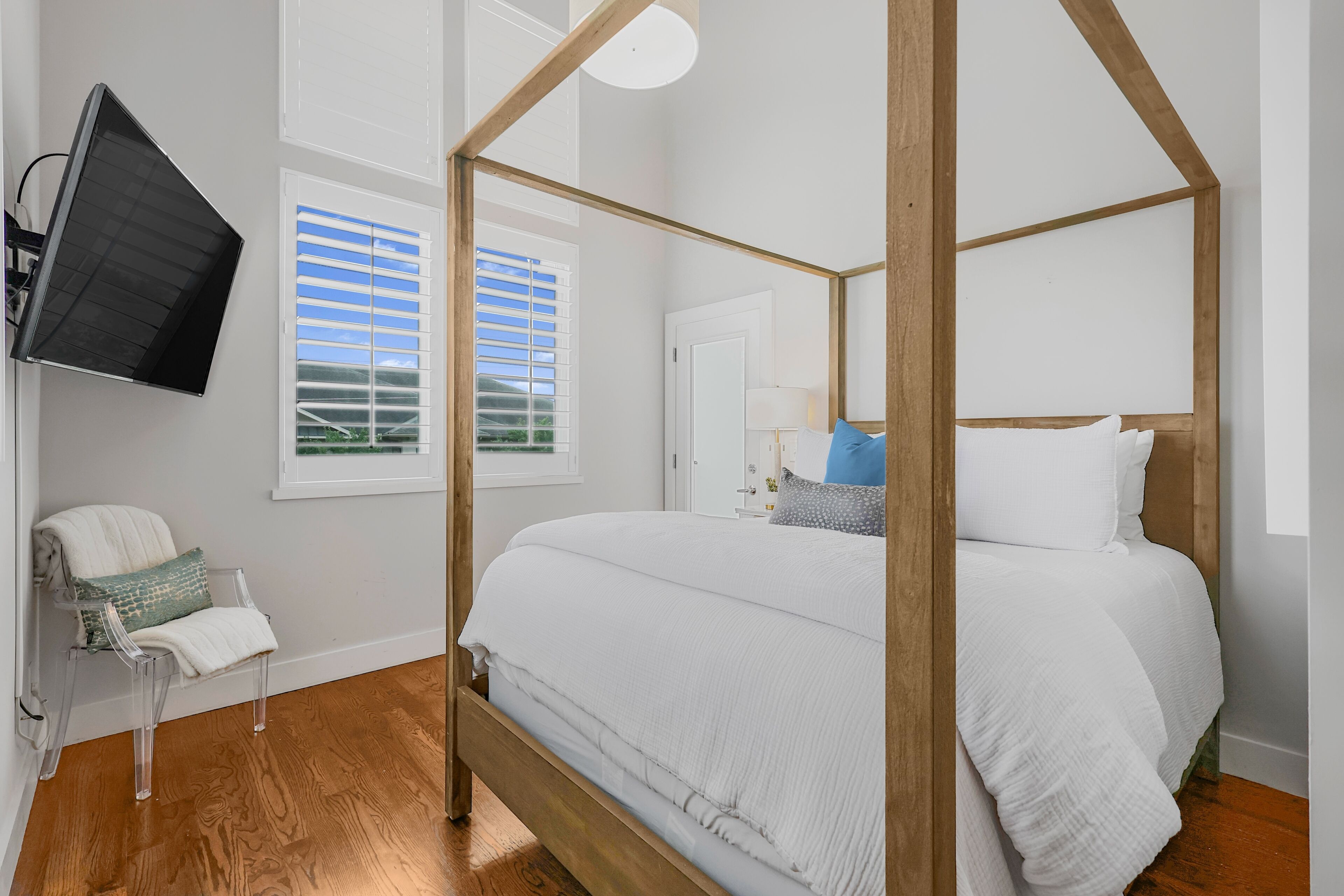 Ein Bett mit Baldachin, ausgestattet mit einer weißen Decke, ein transparenter Stuhl mit weißem Kissen, ein Flachbildfernseher an der Wand und ein Fenster mit Jalousien.