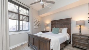 Un dormitorio con una cama grande, un ventilador de techo, una mesita de noche con lámpara y una ventana con cortinas.