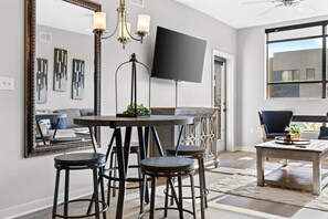 Un salon moderne doté d’un grand téléviseur à écran plat, d’une table de salle à manger ronde avec des tabourets, d’un ventilateur de plafond et d’un grand miroir.