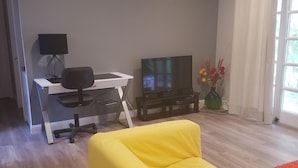 A living room with a yellow sofa, a TV, a desk with a chair, and a window with curtains. - 3 bed 2 bath home in heart of Silicon Valley (Redwood City)