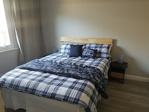 A neatly made bed with a plaid bedspread and pillows, a wooden headboard, a bedside table with a lamp, and a window with curtains.