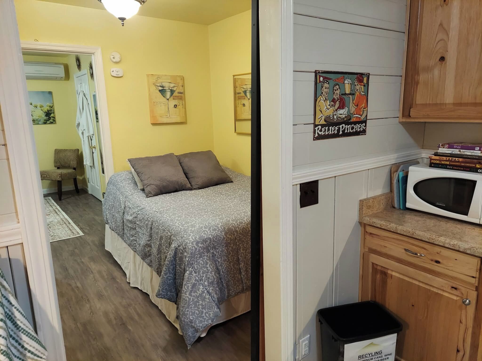 A bedroom with a bed, a small desk, a microwave, and a recycling bin.