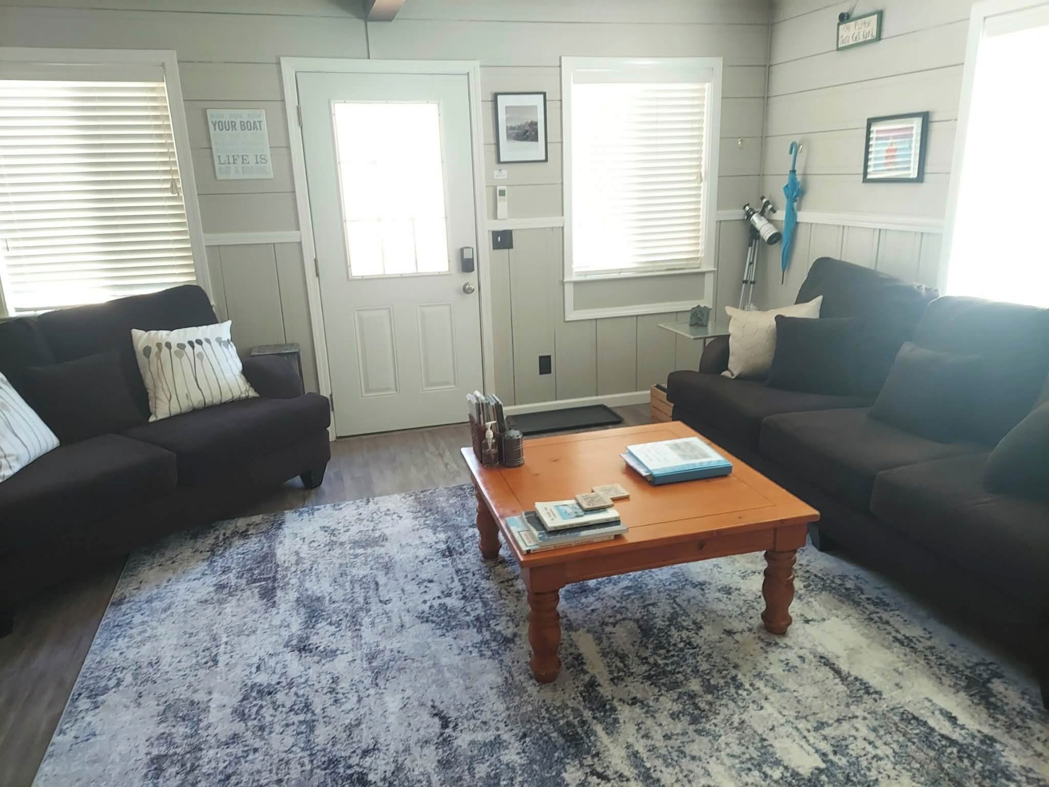 A living room with two dark sofas, a wooden coffee table with books, a blue patterned rug, and a door with a sign.