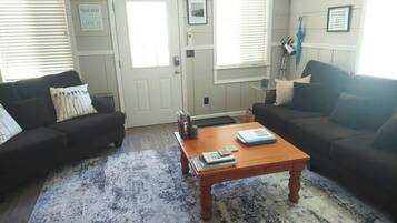 A living room with two dark sofas, a wooden coffee table with books, a blue patterned rug, and a door with a sign.