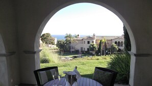 A round table with chairs and a view through an archway to a landscaped area with a building and the ocean in the distance.