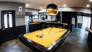 A pool table with a yellow felt surface, surrounded by wooden chairs and a hanging light fixture.