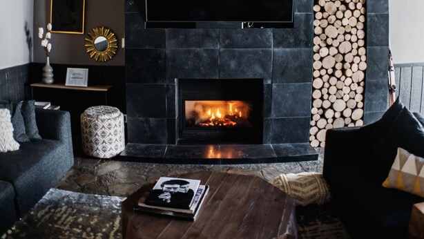 A cozy living room with a stone fireplace, a flat-screen TV mounted above it, and a stack of firewood to the right.